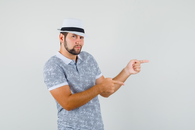 Jonge man wijst opzij in gestreept t-shirt, hoed en ziet er schattig uit. vooraanzicht.