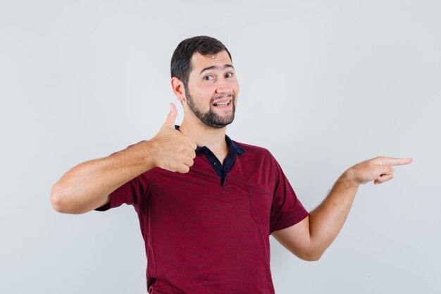 Jonge man wijst opzij terwijl duim omhoog in rood t-shirt en kijkt tevreden, vooraanzicht.