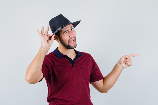 Jonge man wijst opzij terwijl het aanraken van hoed in t-shirt, hoed en op zoek vrolijk. vooraanzicht.