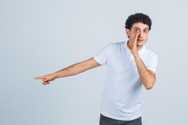 Jonge man wijst opzij, vertelt geheim in wit t-shirt, broek en kijkt nieuwsgierig, vooraanzicht.