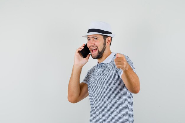 Jonge man wijst tijdens het praten op mobiele telefoon in t-shirt, hoed en ziet er vrolijk uit, vooraanzicht.