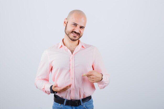 Jonge man wijzend naar zijn hand in roze shirt, spijkerbroek, vooraanzicht.