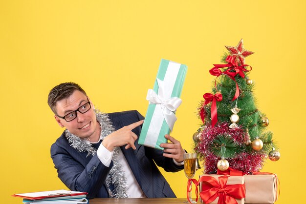 jonge man wijzend op cadeau zitten aan de tafel in de buurt van kerstboom en presenteert op geel