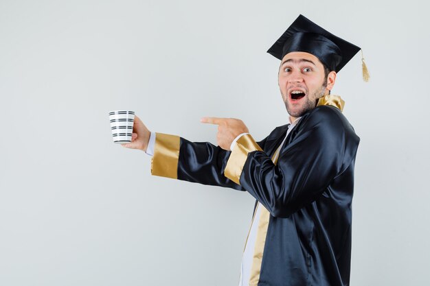 Jonge man wijzend op kopje drank in afgestudeerde uniform en op zoek gelukkig.​