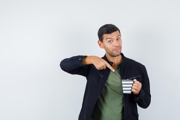 Jonge man wijzend op kopje drank in t-shirt, jas en op zoek gelukkig, vooraanzicht.