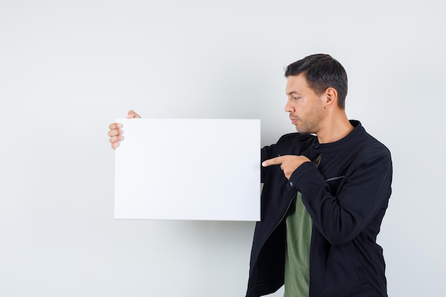 Jonge man wijzend op leeg canvas in t-shirt, jas en gefocust op zoek. vooraanzicht.