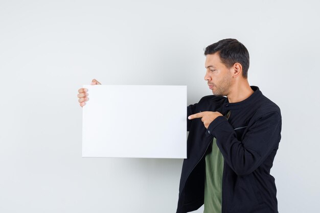 Jonge man wijzend op leeg canvas in t-shirt, jas en gefocust op zoek. vooraanzicht.