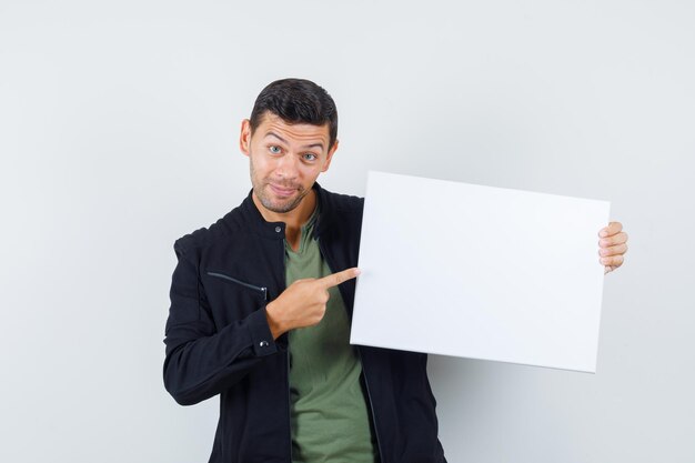 Jonge man wijzend op leeg canvas in t-shirt, jas en vrolijk, vooraanzicht.