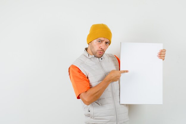 Jonge man wijzend op leeg canvas in t-shirt, jas, hoed vooraanzicht.