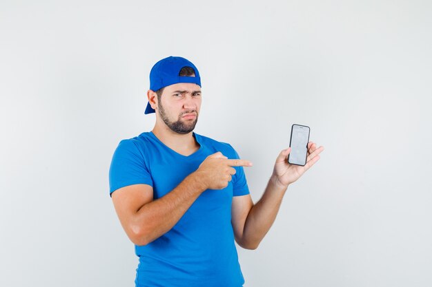 Jonge man wijzend op mobiele telefoon in blauw t-shirt en pet en kijkt ontevreden