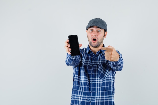 Jonge man wijzend op mobiele telefoon in shirt, pet en kijkt verbaasd, vooraanzicht.