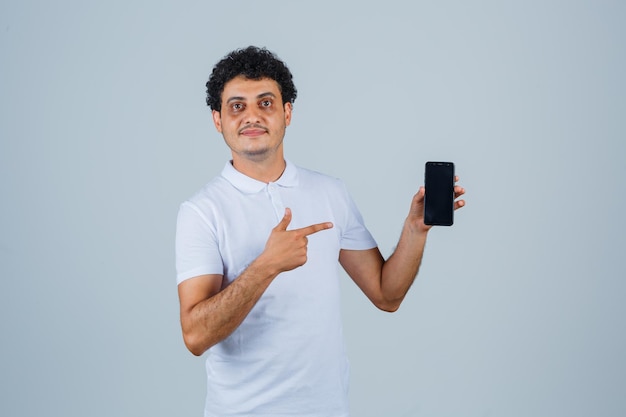 Jonge man wijzend op mobiele telefoon in wit t-shirt en zelfverzekerd kijken. vooraanzicht.