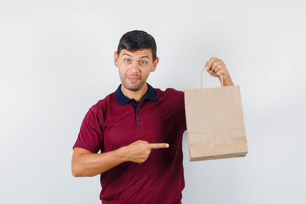 Jonge man wijzend op papieren zak in t-shirt en nieuwsgierig kijkend, vooraanzicht.