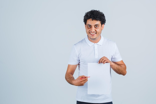 Jonge man wijzend op vel papier met pen in wit t-shirt, broek en zelfverzekerd, vooraanzicht.