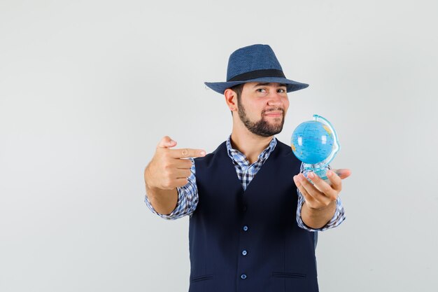 Jonge man wijzend op wereldbol in overhemd, vest, hoed en op zoek zelfverzekerd. vooraanzicht.