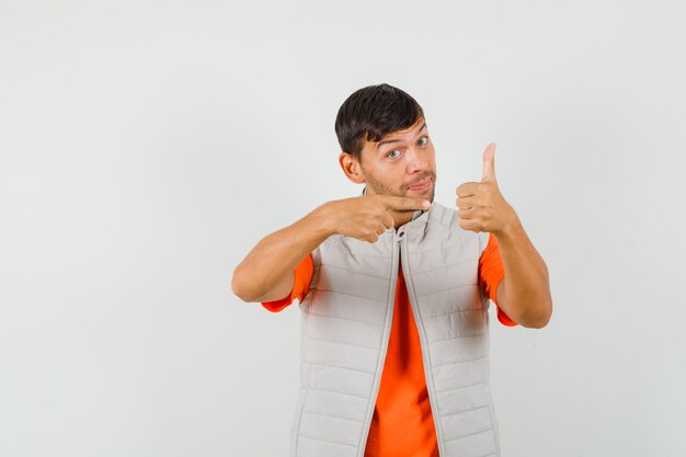 Jonge man wijzend op zijn duim omhoog in t-shirt, jasje en op zoek zelfverzekerd. vooraanzicht.