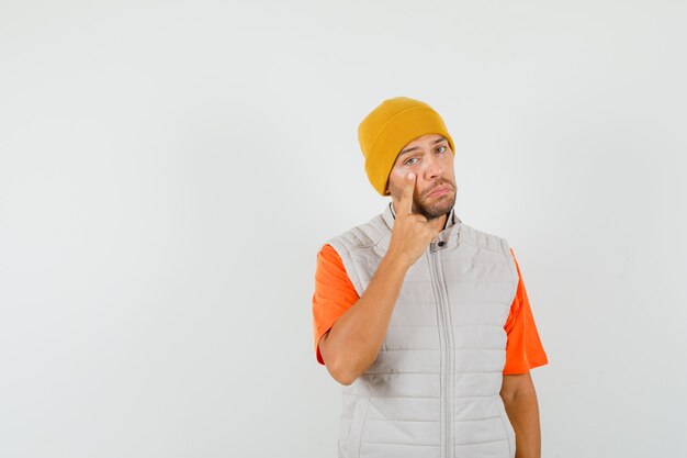 Jonge man wijzend op zijn onderste ooglid in t-shirt, jas, hoed en op zoek verdrietig. vooraanzicht.