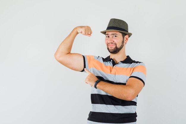 Jonge man wijzend op zijn spieren in t-shirt, hoed en op zoek zelfverzekerd. vooraanzicht.