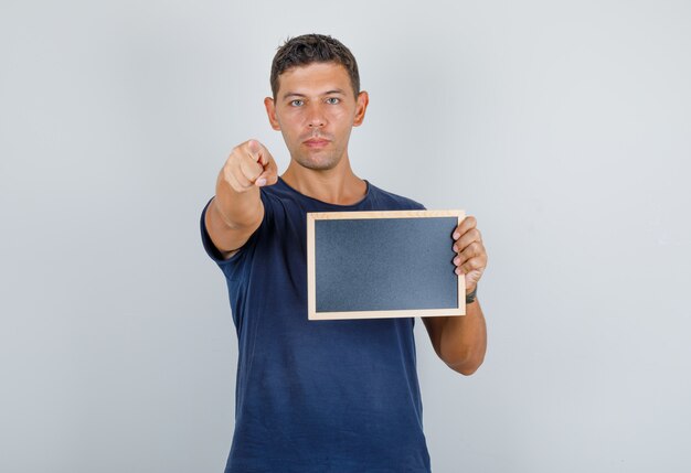 Jonge man wijzende vinger naar camera met bord in donkerblauw t-shirt, vooraanzicht.