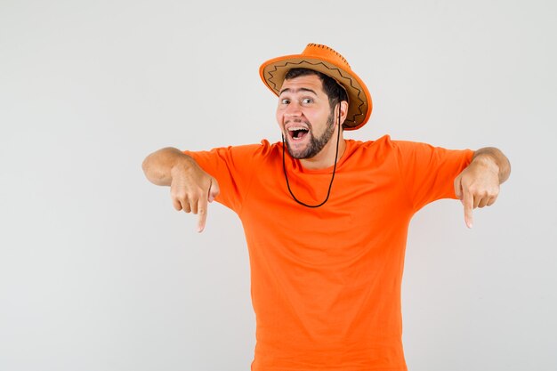 Jonge man wijzende vingers naar beneden in oranje t-shirt, hoed en op zoek vrolijk, vooraanzicht.
