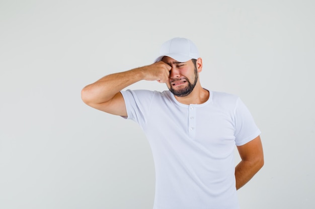 Jonge man zijn ogen wrijven tijdens het huilen in t-shirt, pet en op zoek verdrietig. vooraanzicht.