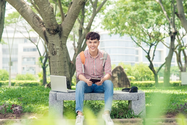 Jonge man zittend op een bankje in het park, boek lezen, glimlachen en kijken naar de camera