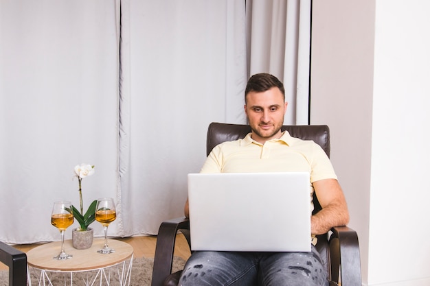 Gratis foto jonge man zittend op een stoel met behulp van laptop met wijnglazen op tafel