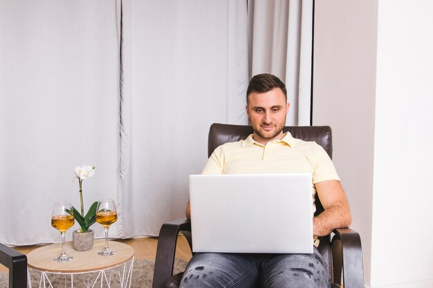 Jonge man zittend op een stoel met behulp van laptop met wijnglazen op tafel