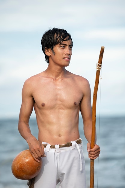 Gratis foto jonge man zonder shirt op het strand met houten strik die zich voorbereidt om capoeira . te oefenen