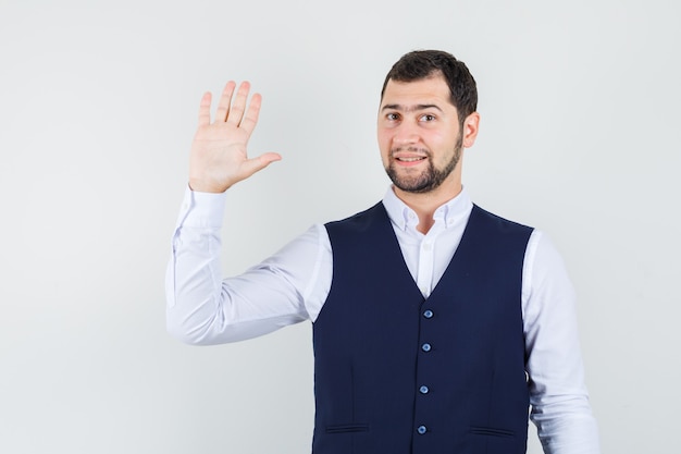 Jonge man zwaaiende hand voor begroeting in overhemd en vest en op zoek vrolijk