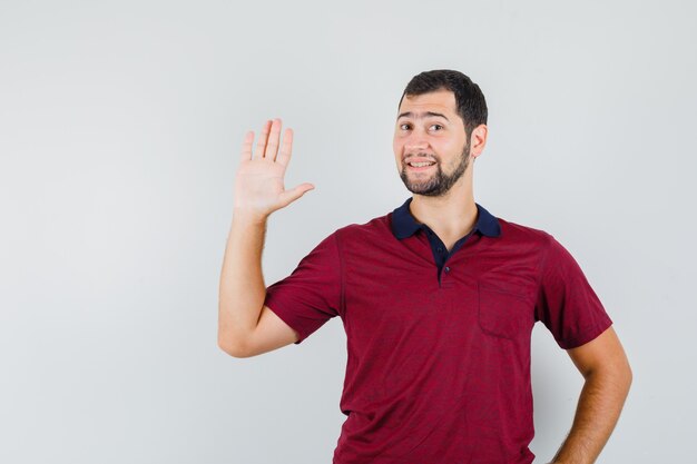 Jonge man zwaaiende hand voor groet in rood t-shirt en op zoek optimistisch. vooraanzicht.