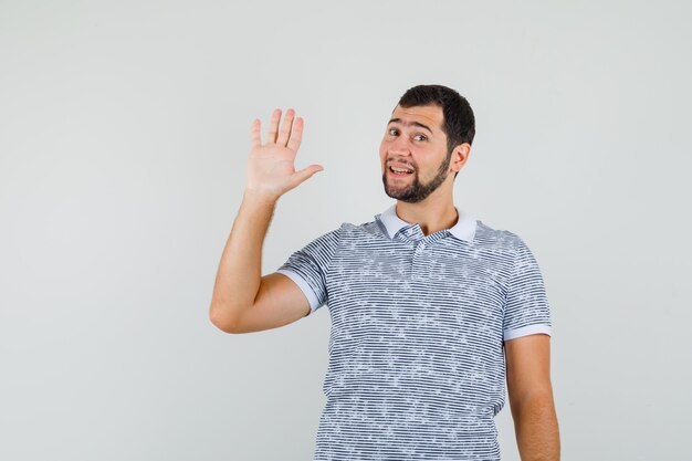 Jonge man zwaaiende hand voor groet in t-shirt en op zoek vrolijk, vooraanzicht.