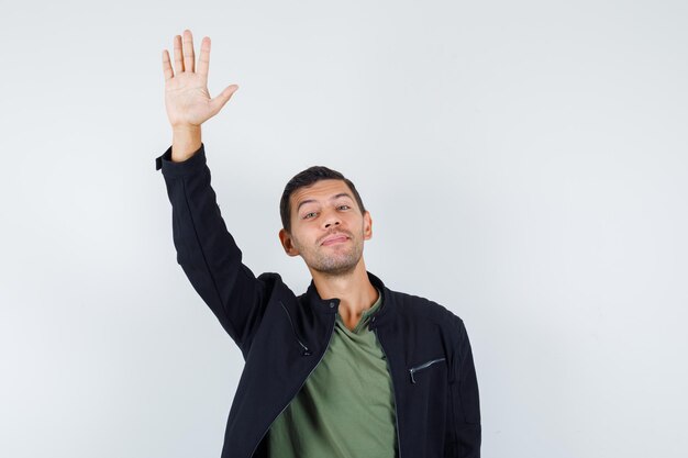 Jonge man zwaait met de hand voor begroeting in t-shirt, jas en ziet er vrolijk uit, vooraanzicht.