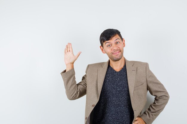 Jonge man zwaait met zijn hand voor afscheid in grijsachtig bruin jasje, zwart hemd vooraanzicht.