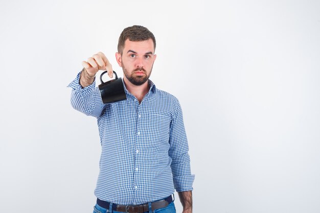 Jonge mannelijke arm uitrekken terwijl handvat van beker in shirt, spijkerbroek en op zoek zelfverzekerd. vooraanzicht.