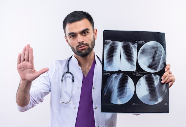 jonge mannelijke arts die stethoscoop medische toga draagt die x-ray houdt die stopgebaar op geïsoleerde witte muur toont