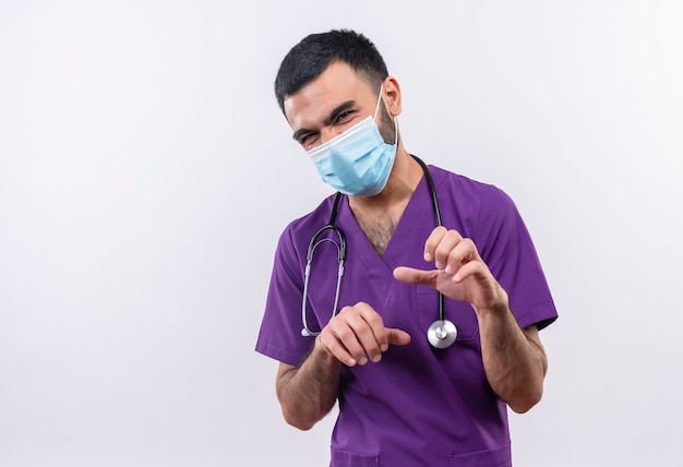 jonge mannelijke arts draagt paarse chirurg kleding en stethoscoop medisch masker met tijger gebaar op geïsoleerde witte muur