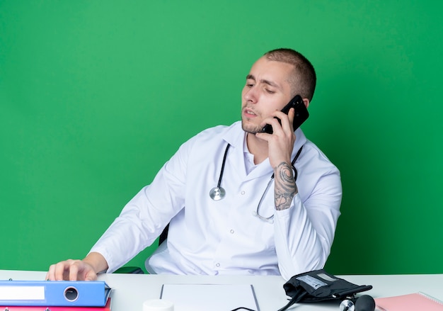 Jonge mannelijke arts dragen medische gewaad en stethoscoop zittend aan een bureau met werkgereedschap praten over de telefoon en neerkijkt met de hand op Bureau geïsoleerd op groene achtergrond