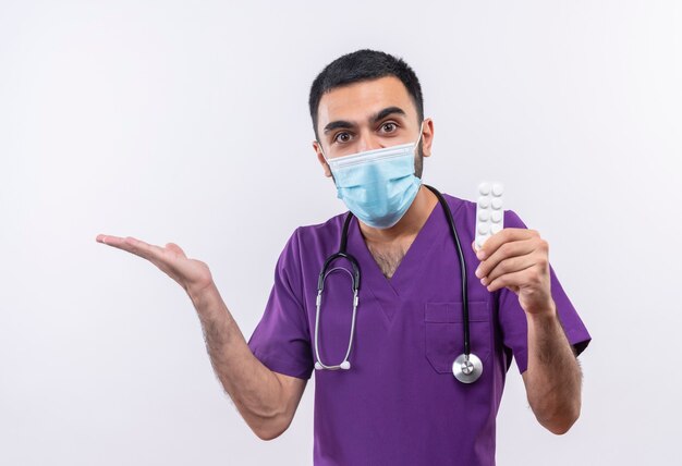 jonge mannelijke arts dragen paarse chirurg kleding en stethoscoop medisch masker pillen op geïsoleerde witte muur te houden
