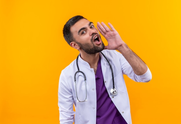 Jonge mannelijke arts met stethoscoop medische jurk fluistert op geïsoleerde gele muur