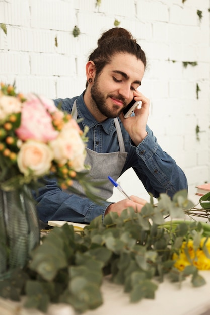 Gratis foto jonge mannelijke bloemist die cliëntorde opmerkt tijdens mobiel telefoongesprek in bloemwinkel