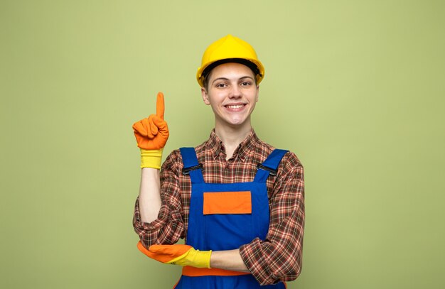 Jonge mannelijke bouwer die uniform draagt met handschoenen geïsoleerd op olijfgroene muur