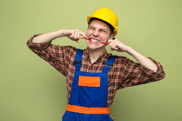 Gratis foto jonge mannelijke bouwer verzegelde mond met ducttape in uniform geïsoleerd op olijfgroene muur