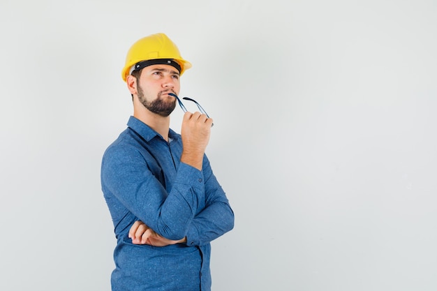 Jonge mannelijke bril bijten tijdens het kijken in shirt, helm en peinzend kijken. vooraanzicht.