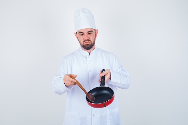 Jonge mannelijke chef-kok die in wit uniform lege koekenpan met houten lepel houdt en terneergeslagen, vooraanzicht kijkt.