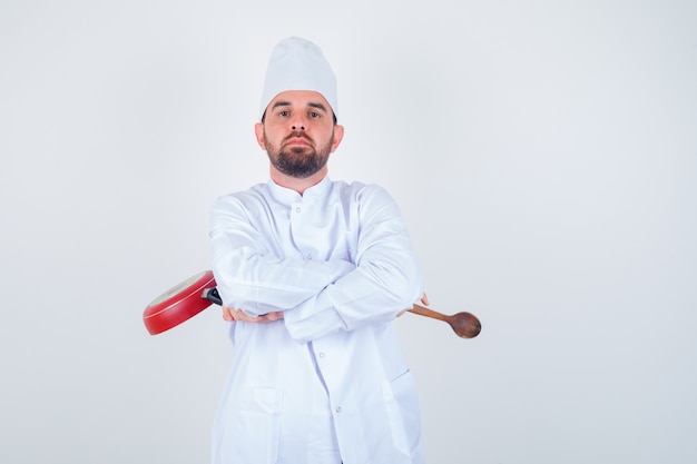 Jonge mannelijke chef-kok met koekenpan en houten lepel terwijl hij met gekruiste armen in wit uniform staat en er zelfverzekerd uitziet, vooraanzicht.