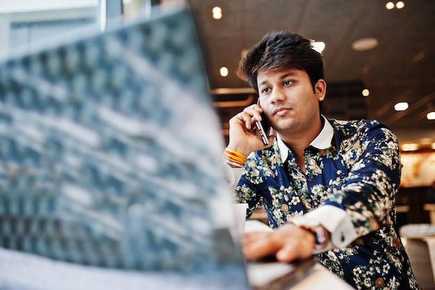 Gratis foto jonge mannelijke indiase freelancer zit met opengeklapte laptopcomputer in fastfoodcafé knappe aziatische man die telefoon spreekt tijdens het werk op netbook in comfortabele coffeeshop