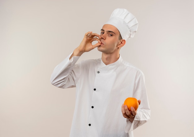 jonge mannelijke kok die chef-kok uniform draagt die sinaasappel toont die heerlijk gebaar op geïsoleerde witte muur met exemplaarruimte toont