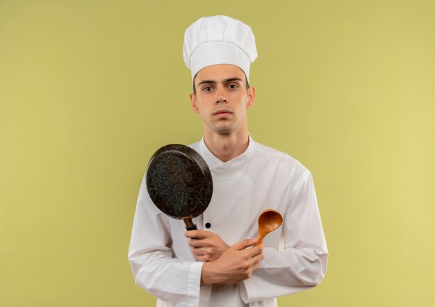 jonge mannelijke kok dragen uniforme chef-kok kruising koekenpan en lepel in zijn hand op geïsoleerde groene muur met kopie ruimte