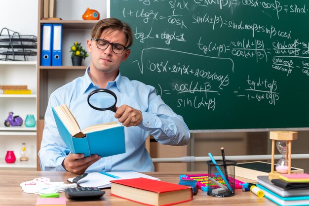 Jonge mannelijke leraar bril zit op school bureau met boeken en notities kijken door vergrootglas naar boek met ernstig gezicht voor schoolbord in klas